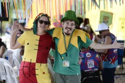 Bloco Deficiente é a Mãe - Duas pessoas fantasiadas e sorrindo, com os braços abertos, em um ambiente festivo decorado com fitas coloridas. Ao fundo, outras pessoas participam da celebração, algumas sentadas em cadeiras plásticas.