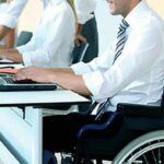 Homem em cadeira de rodas trabalhando em um escritório, digitando em um notebook sobre a mesa, enquanto outros colegas também utilizam computadores ao fundo.