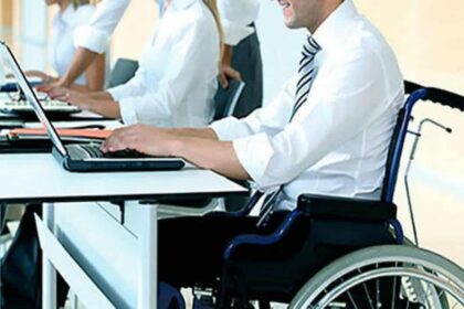 Homem em cadeira de rodas trabalhando em um escritório, digitando em um notebook sobre a mesa, enquanto outros colegas também utilizam computadores ao fundo.