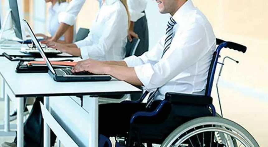 Homem em cadeira de rodas trabalhando em um escritório, digitando em um notebook sobre a mesa, enquanto outros colegas também utilizam computadores ao fundo.