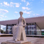 Estátua da Justiça em frente ao prédio do Supremo Tribunal Federal em Brasília, com céu azul ao fundo.