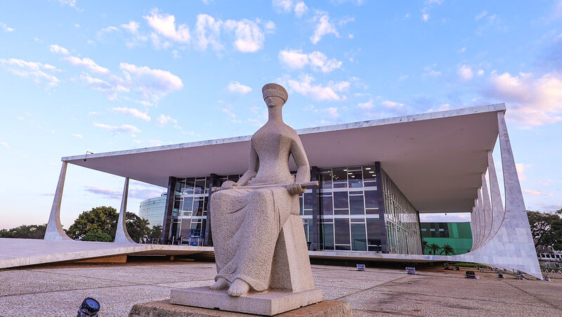 Estátua da Justiça em frente ao prédio do Supremo Tribunal Federal em Brasília, com céu azul ao fundo.