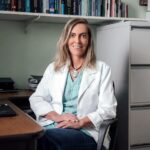 Tatiana Coelho vestindo jaleco branco sentada em um consultório, sorrindo para a câmera. Ao fundo, há estante com livros, armário de arquivos e mesa com computador, indicando ambiente profissional da área da saúde.