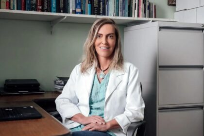 Tatiana Coelho vestindo jaleco branco sentada em um consultório, sorrindo para a câmera. Ao fundo, há estante com livros, armário de arquivos e mesa com computador, indicando ambiente profissional da área da saúde.