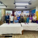 Grupo de pessoas reunidas em uma sala após evento institucional, posicionadas atrás de duas mesas cobertas por toalhas brancas. Ao fundo, banners de organizações e materiais informativos relacionados a políticas públicas e direitos das pessoas com deficiência.