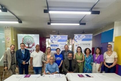 Grupo de pessoas reunidas em uma sala após evento institucional, posicionadas atrás de duas mesas cobertas por toalhas brancas. Ao fundo, banners de organizações e materiais informativos relacionados a políticas públicas e direitos das pessoas com deficiência.