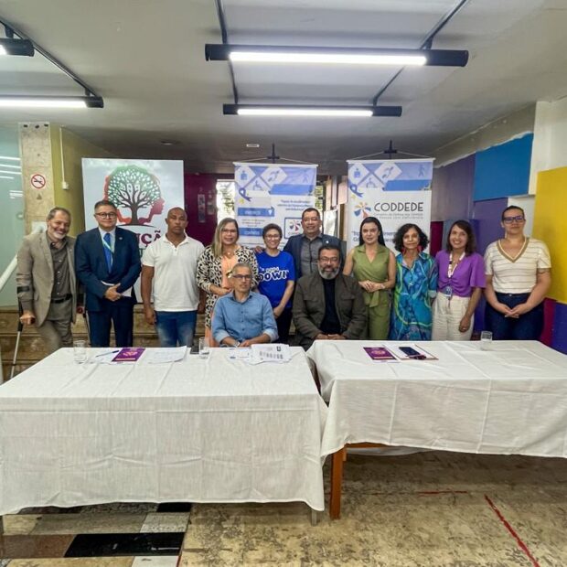 Grupo de pessoas reunidas em uma sala após evento institucional, posicionadas atrás de duas mesas cobertas por toalhas brancas. Ao fundo, banners de organizações e materiais informativos relacionados a políticas públicas e direitos das pessoas com deficiência.