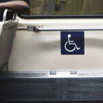 Interior de transporte público com assento preferencial sinalizado pelo símbolo de acessibilidade. Ao lado, uma pessoa sentada usa fones de ouvido, enquanto a área reservada para pessoas com deficiência está destacada na parede.