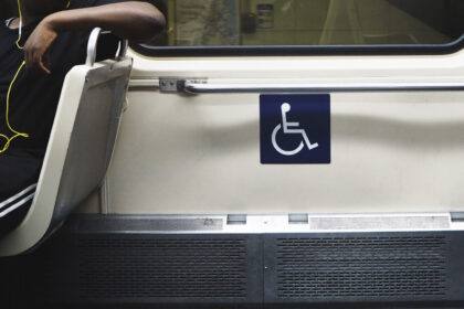 Interior de transporte público com assento preferencial sinalizado pelo símbolo de acessibilidade. Ao lado, uma pessoa sentada usa fones de ouvido, enquanto a área reservada para pessoas com deficiência está destacada na parede.