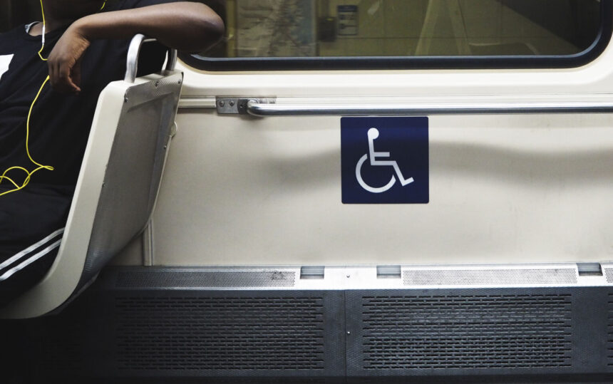Interior de transporte público com assento preferencial sinalizado pelo símbolo de acessibilidade. Ao lado, uma pessoa sentada usa fones de ouvido, enquanto a área reservada para pessoas com deficiência está destacada na parede.