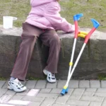 Criança sentada em um banco de pedra ao ar livre, usando casaco rosa, com um par de muletas apoiadas ao lado. No chão há desenhos de giz e ao fundo aparece uma área gramada. Adoção de crianças com deficiência no Brasil
