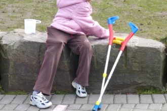 Criança sentada em um banco de pedra ao ar livre, usando casaco rosa, com um par de muletas apoiadas ao lado. No chão há desenhos de giz e ao fundo aparece uma área gramada. Adoção de crianças com deficiência no Brasil
