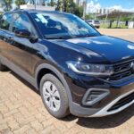 SUV T-cross compacto preto estacionado em área externa, com design moderno, faróis afilados e grade frontal com logotipo da Volkswagen. O veículo está em um pátio pavimentado, com outros carros e prédios ao fundo.