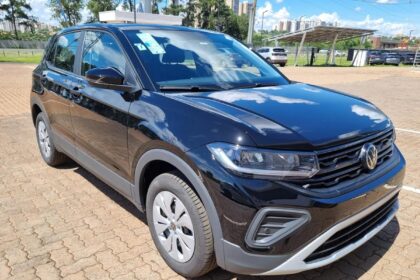 SUV T-cross compacto preto estacionado em área externa, com design moderno, faróis afilados e grade frontal com logotipo da Volkswagen. O veículo está em um pátio pavimentado, com outros carros e prédios ao fundo.