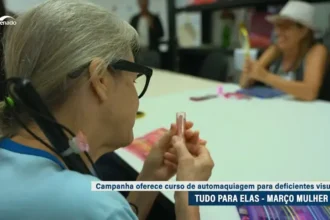 Mulher com deficiência visual sentada à mesa, segurando um batom durante atividade de automaquiagem. Ao fundo, outra mulher também participa da oficina. Na tela, aparece a legenda: “Campanha oferece curso de automaquiagem para deficientes visuais – Tudo para elas – Março Mulher”, com logotipo da TV Senado no canto superior.