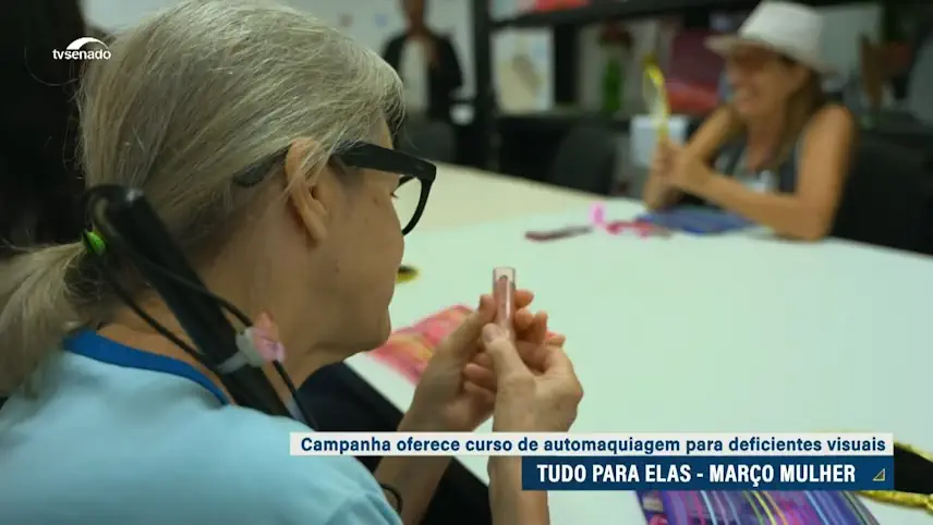 Mulher com deficiência visual sentada à mesa, segurando um batom durante atividade de automaquiagem. Ao fundo, outra mulher também participa da oficina. Na tela, aparece a legenda: “Campanha oferece curso de automaquiagem para deficientes visuais – Tudo para elas – Março Mulher”, com logotipo da TV Senado no canto superior.