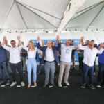 Grupo de autoridades e participantes em evento público, de mãos dadas e erguidas, sobre um palco sob tenda. Entre eles, há uma pessoa em cadeira de rodas. Ao fundo, banners institucionais indicam ação governamental ou anúncio oficial.