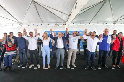 Grupo de autoridades e participantes em evento público, de mãos dadas e erguidas, sobre um palco sob tenda. Entre eles, há uma pessoa em cadeira de rodas. Ao fundo, banners institucionais indicam ação governamental ou anúncio oficial.