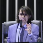 Pessoa falando ao microfone em uma sessão formal, vestindo terno e óculos, sentada em uma cadeira de destaque. Ao fundo, aparecem elementos de um plenário ou auditório institucional.