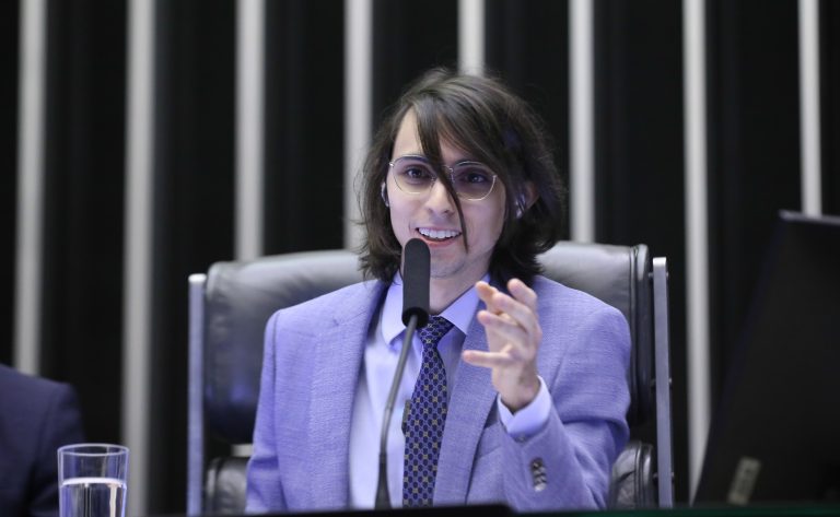 Pessoa falando ao microfone em uma sessão formal, vestindo terno e óculos, sentada em uma cadeira de destaque. Ao fundo, aparecem elementos de um plenário ou auditório institucional.