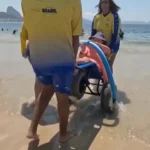 Pessoa com deficiência sendo conduzida por monitores em uma cadeira anfíbia na água do mar, durante atividade de acessibilidade na praia. Outras pessoas e banhistas aparecem ao fundo, enquanto a equipe utiliza camisetas amarelas de identificação.