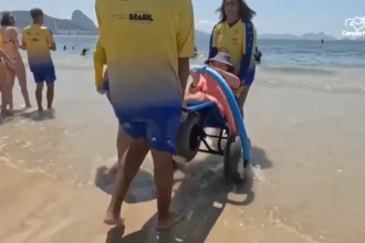 Pessoa com deficiência sendo conduzida por monitores em uma cadeira anfíbia na água do mar, durante atividade de acessibilidade na praia. Outras pessoas e banhistas aparecem ao fundo, enquanto a equipe utiliza camisetas amarelas de identificação.