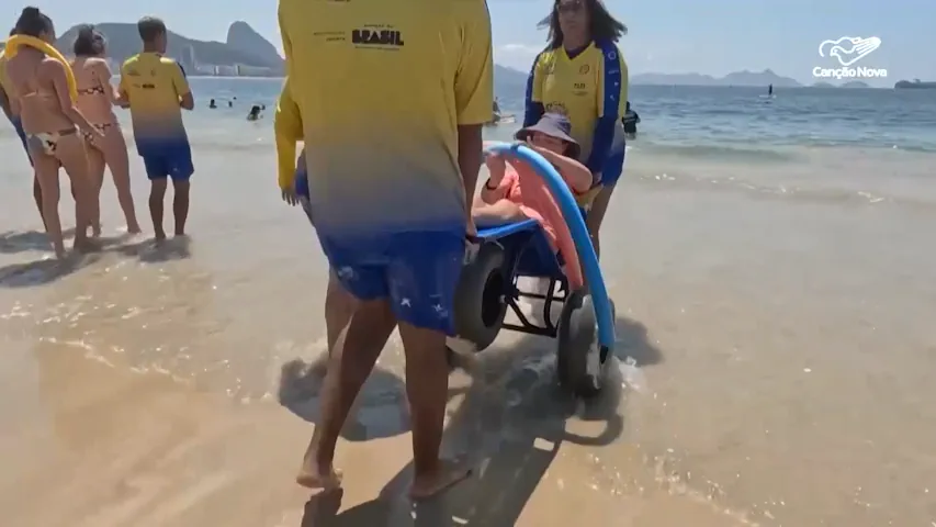 Pessoa com deficiência sendo conduzida por monitores em uma cadeira anfíbia na água do mar, durante atividade de acessibilidade na praia. Outras pessoas e banhistas aparecem ao fundo, enquanto a equipe utiliza camisetas amarelas de identificação.