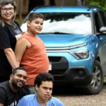 Família posando ao ar livre em frente a um carro azul estacionado. Duas pessoas estão em pé e duas agachadas à frente, sorrindo para a câmera, em ambiente com árvores e vegetação ao fundo.