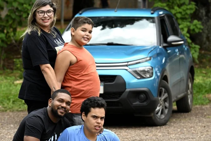 Família posando ao ar livre em frente a um carro azul estacionado. Duas pessoas estão em pé e duas agachadas à frente, sorrindo para a câmera, em ambiente com árvores e vegetação ao fundo.