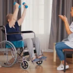 Pessoa idosa em cadeira de rodas realizando exercícios com halteres, levantando os braços, enquanto uma profissional de saúde sentada à frente orienta e acompanha a atividade em ambiente interno, representando reabilitação e atividade física inclusiva.