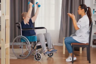 Pessoa idosa em cadeira de rodas realizando exercícios com halteres, levantando os braços, enquanto uma profissional de saúde sentada à frente orienta e acompanha a atividade em ambiente interno, representando reabilitação e atividade física inclusiva.