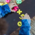 Mãos de uma criança manipulando letras e números coloridos de plástico sobre uma superfície escura, em uma mesa de madeira, representando atividades de aprendizado e educação inclusiva.