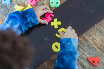 Mãos de uma criança manipulando letras e números coloridos de plástico sobre uma superfície escura, em uma mesa de madeira, representando atividades de aprendizado e educação inclusiva.