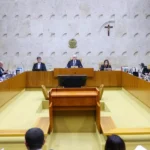 A imagem mostra uma sessão de julgamento no Supremo Tribunal Federal (STF). No centro, há vários ministros sentados em uma bancada em formato semicircular, conduzindo uma audiência formal. Ao fundo, é possível ver a bandeira do Brasil e um crucifixo na parede — elementos típicos do plenário do STF. Também aparecem telas exibindo a transmissão da sessão, indicando que o julgamento está sendo acompanhado ao vivo.