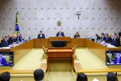 A imagem mostra uma sessão de julgamento no Supremo Tribunal Federal (STF). No centro, há vários ministros sentados em uma bancada em formato semicircular, conduzindo uma audiência formal. Ao fundo, é possível ver a bandeira do Brasil e um crucifixo na parede — elementos típicos do plenário do STF. Também aparecem telas exibindo a transmissão da sessão, indicando que o julgamento está sendo acompanhado ao vivo.