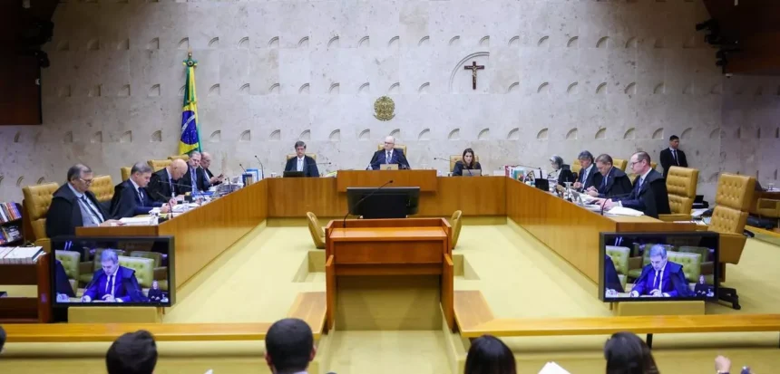 A imagem mostra uma sessão de julgamento no Supremo Tribunal Federal (STF). No centro, há vários ministros sentados em uma bancada em formato semicircular, conduzindo uma audiência formal. Ao fundo, é possível ver a bandeira do Brasil e um crucifixo na parede — elementos típicos do plenário do STF. Também aparecem telas exibindo a transmissão da sessão, indicando que o julgamento está sendo acompanhado ao vivo.