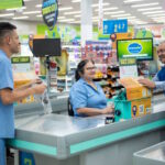 Funcionários atendem clientes em um supermercado, com destaque para uma operadora de caixa sorrindo enquanto realiza o atendimento. O ambiente é organizado, com sinalizações promocionais e produtos ao fundo
