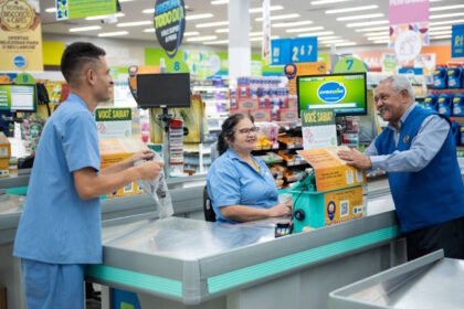 Funcionários atendem clientes em um supermercado, com destaque para uma operadora de caixa sorrindo enquanto realiza o atendimento. O ambiente é organizado, com sinalizações promocionais e produtos ao fundo
