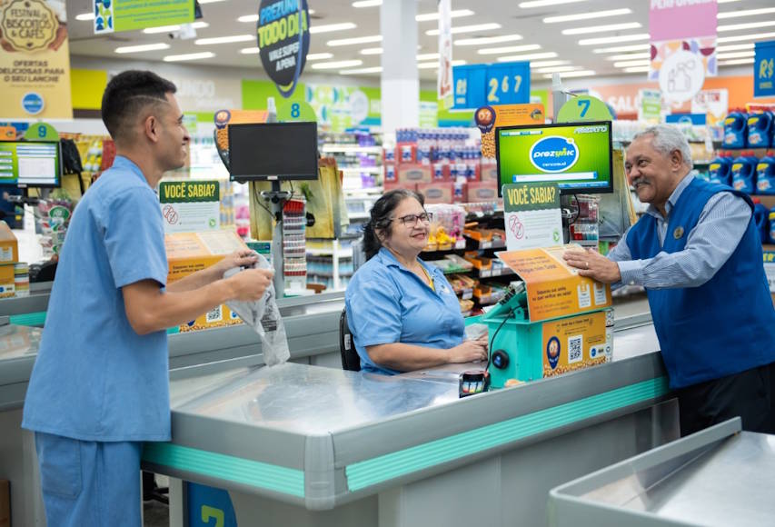 Funcionários atendem clientes em um supermercado, com destaque para uma operadora de caixa sorrindo enquanto realiza o atendimento. O ambiente é organizado, com sinalizações promocionais e produtos ao fundo