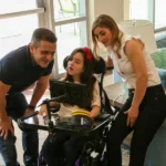 Clara em cadeira de rodas utilizando um dispositivo de comunicação assistiva com tela, enquanto sus pais se inclinam ao lado, interagindo e acompanhando a atividade em um ambiente doméstico.