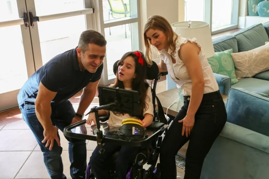 Clara em cadeira de rodas utilizando um dispositivo de comunicação assistiva com tela, enquanto sus pais se inclinam ao lado, interagindo e acompanhando a atividade em um ambiente doméstico.