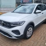 Imagem de um SUV branco Volkswagen T-Cross estacionado em área pavimentada, em frente a um prédio comercial com rampa de acessibilidade e corrimãos. O veículo aparece em destaque, visto de frente e lateralmente.