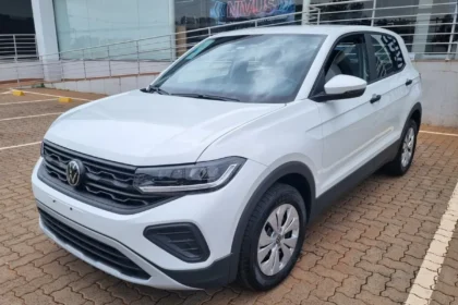 Imagem de um SUV branco Volkswagen T-Cross estacionado em área pavimentada, em frente a um prédio comercial com rampa de acessibilidade e corrimãos. O veículo aparece em destaque, visto de frente e lateralmente.
