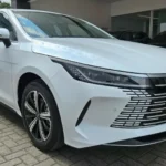 Sedã branco BYD King estacionado em frente a uma concessionária, visto em ângulo frontal e lateral. O carro possui faróis estreitos, grade dianteira moderna com detalhes cromados e rodas esportivas pretas e prata.