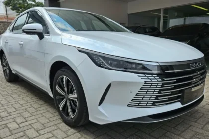 Sedã branco BYD King estacionado em frente a uma concessionária, visto em ângulo frontal e lateral. O carro possui faróis estreitos, grade dianteira moderna com detalhes cromados e rodas esportivas pretas e prata.