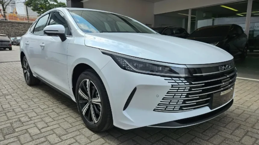 Sedã branco BYD King estacionado em frente a uma concessionária, visto em ângulo frontal e lateral. O carro possui faróis estreitos, grade dianteira moderna com detalhes cromados e rodas esportivas pretas e prata.