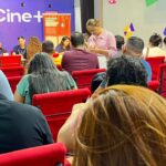 **Texto alternativo (acessibilidade):** Sala de atendimento com várias pessoas sentadas em cadeiras vermelhas, organizadas em fileiras, aguardando atendimento. À frente, há uma mesa com atendentes conversando e registrando informações. Uma mulher em pé preenche um formulário. No fundo, uma parede colorida exibe a palavra “Cine+” e cartazes informativos. Ambiente interno, iluminado e com clima de espera e organização.