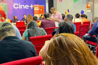**Texto alternativo (acessibilidade):** Sala de atendimento com várias pessoas sentadas em cadeiras vermelhas, organizadas em fileiras, aguardando atendimento. À frente, há uma mesa com atendentes conversando e registrando informações. Uma mulher em pé preenche um formulário. No fundo, uma parede colorida exibe a palavra “Cine+” e cartazes informativos. Ambiente interno, iluminado e com clima de espera e organização.