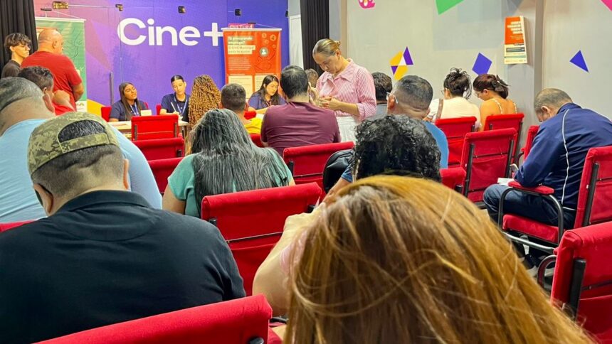 **Texto alternativo (acessibilidade):** Sala de atendimento com várias pessoas sentadas em cadeiras vermelhas, organizadas em fileiras, aguardando atendimento. À frente, há uma mesa com atendentes conversando e registrando informações. Uma mulher em pé preenche um formulário. No fundo, uma parede colorida exibe a palavra “Cine+” e cartazes informativos. Ambiente interno, iluminado e com clima de espera e organização.