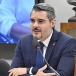Homem de terno e gravata falando ao microfone durante uma reunião ou audiência pública, sentado à mesa, com expressão séria e gesticulando com as mãos. Ao fundo, há uma tela e elementos de um ambiente institucional.